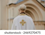 Back view medium shot of unrecognizable senior Catholic priest standing outdoors in front of church building, copy space