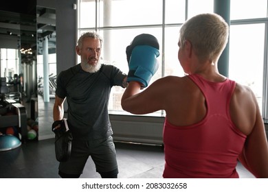 Back View Middle Age Woman With Short Blond Hair Wearing Pink Top And Blue Boxing Gloves Hits Trainer Into Punch Mits. Mature Professional Trainer Helps Woman To Learn Boxing Techniques.