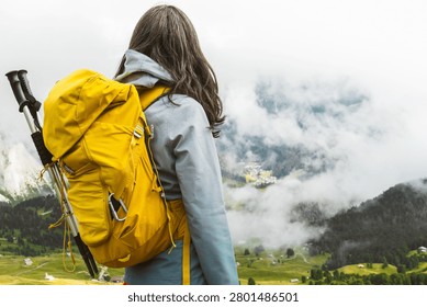 Back view of middle aged gray hair woman hiker yellow backpack overlooking lush green alpine valley misty mountains background Dolomites Alps Italy summer landscape.