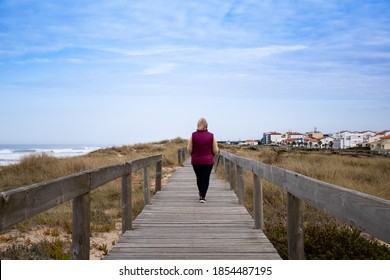 Back view of middle-aged woman walking alone on boardwalk at the beach and distant houses. Autumn walk by the sea near the neighborhood. Health, social distancing and retirement concepts - Powered by Shutterstock