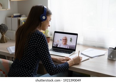 Back view of millennial female student in headphones note study distant with teacher or tutor on laptop. Young woman in earphones watch webinar, have online training or course with coach on computer.