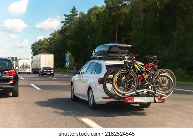 Back view modern white family wagon car with mounted roof box trunk and bike tail carrier driving european highway road against blue sky summer day. Lifestyle travel adventure trip journey concept - Shutterstock ID 2182524545