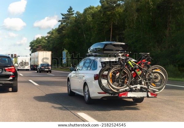 Back view modern white family wagon car with\
mounted roof box trunk and bike tail carrier driving european\
highway road against blue sky summer day. Lifestyle travel\
adventure trip journey\
concept