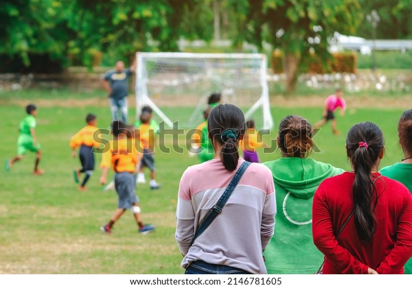 Back view of Moms watch and cheering their sons
playing football in school tournament on sideline. Sport, outdoor
active,  Spectator watching soccer game. Parents care and encourage
their children.