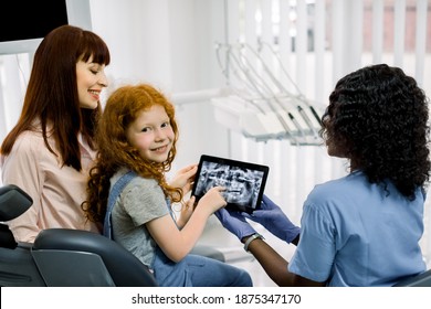 Back View Of Mother And Child Girl At Appointment With Female Black Doctor Orthodontist, Showing Xray Teeth Scan On Tablet. Dental Examination And Dentist Consultation. Little Girl Looks At Camera