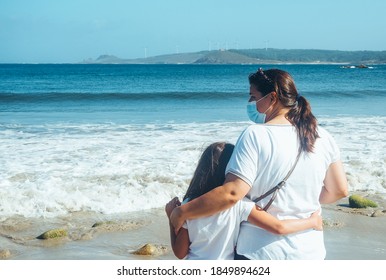 Back View Of A Mother And Daughter Embracing With Coronavirus Face Masks Looking At The Sea. New Normal Concept