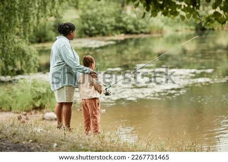 Back view of mother and daughter fishing together by lake in beautiful family bonding moment, copy space 