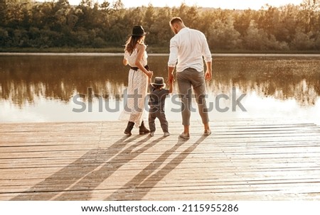 Back view of mother and father holding hands of son while standing on wooden quay near calm water of pond at sunset