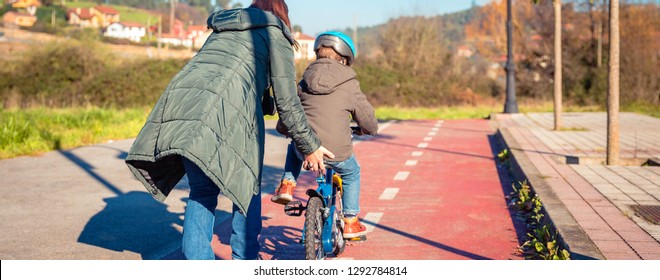 Back View Of Mother Teaching Her Son To Ride A Bicycles In The City On A Sunny Winter Day.  Family Leisure Outdoors Concept.