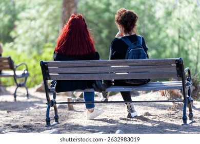 Back view of multi ethnic two ladies sitting on wooden bench in friendship while enjoying the scenic view of peaceful green park.