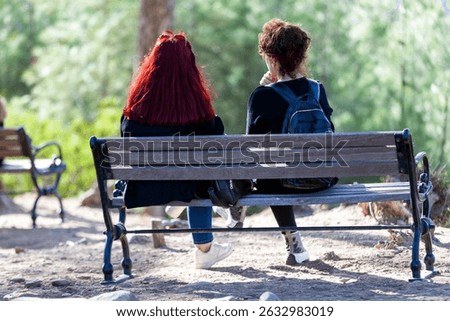 Back view of multi ethnic two ladies sitting on wooden bench in friendship while enjoying the scenic view of peaceful green park.