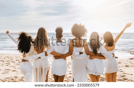 Back view of multiracial ladies in white dresses posing on beach and looking at ocean, having maiden evening, hen party outdoors. Group of women embracing