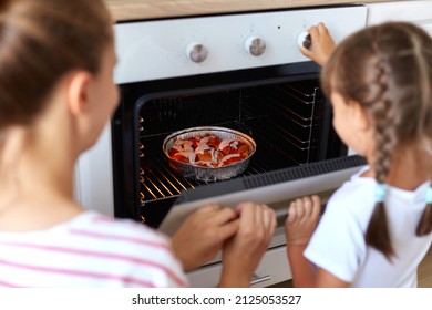 Back view of mum and kid daughter bake muffins in kitchen, mother teaching child take tray with cupcakes dessert, make cakes out oven stove, cook at home, family bakery.