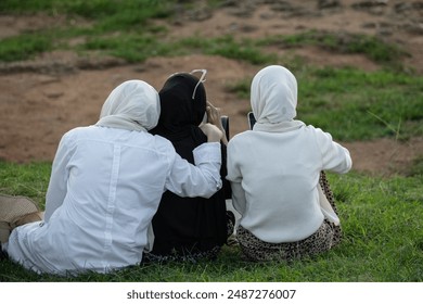 Back view of the muslim women relaxing on green grass in the nature. Three Unrecognizable Muslim Ladies In Traditional Hijab Headscarf In City Park Outdoors. Female Friendship concept - Powered by Shutterstock