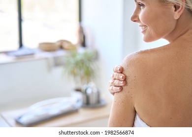 Back View Of Nude Beautiful Middle Aged 50s Woman Applying Exfoliating Peeling Sugar Scrub After Shower. Advertising Of Bodycare Professional Spa Therapy At Home Using Antiage Bodycare Products.