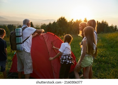 Back View Of Old Man With Rucksack Setting Up Red Tent Together With Kids In Hills. Classmates Organizing Campsite With Teacher With Gray Hair. Concept Of Hiking.