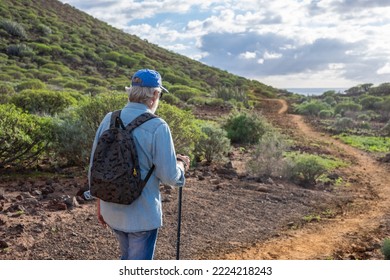 Back view of old senior man enjoying trekking in countryside along the sea. Elderly caucasian male with hat and backpack walking in footpath - Shutterstock ID 2224218243