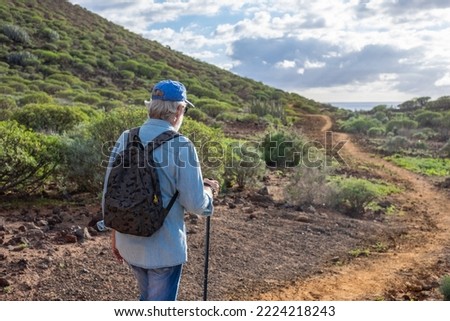 Back view of old senior man enjoying trekking in countryside along the sea. Elderly caucasian male with hat and backpack walking in footpath