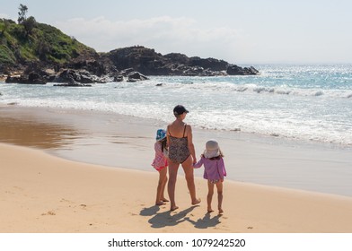 Back View Of Older Woman And Young Girls In Swimming Costumes Walking Towards Water At Beach - Grandparent, Grandchildren Concept