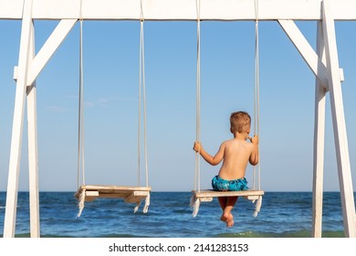 Back view of on alone cute little adorable small caucasian sad pensive kid boy swinging and playing at playground sea sand beach against blue ocean water wave and clear sky. Family vacation - Powered by Shutterstock