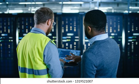 Back View on Bearded IT Engineer in Glasses and High Visibility Vest with a Laptop Computer and Black Technician Colleague Talking in Data Center while Standing Next to Server Racks. - Powered by Shutterstock