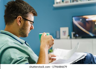 Back View On Caucasian Man Sitting On The Sofa Couch At Home In Front TV Holding A Cup Of Tea Or Coffee Reading Reports Documents Or Analyzing Invoice Bills - Work From Home Concept Real People