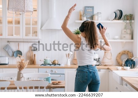 Back view on dancing beautiful Italian girl in headphones, white vest and blue denim jeans listening woman rises hand holds phone. Playful young hispanic woman enjoys weekend at home, kitchen.