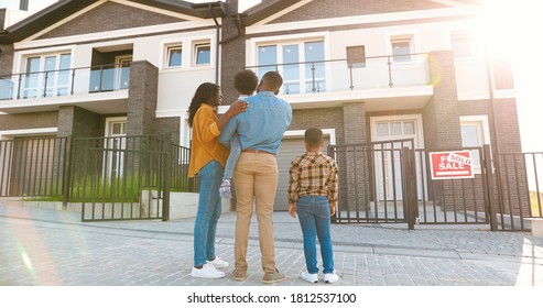 Back View On Happy African American Family With Small Children Standing Near New House At Suburb And Looking On It. 