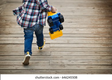 Back View On Little Toddler Boy With His Toy Tractor In His Hands Running On Wooden Bridge Outdoors. Child Rushing Away. Lifestyle