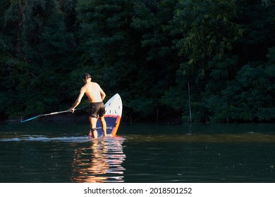 Back view on man making pivot or step back turn trick on stand-up paddle board (SUP) on the river near tress at suumer sunset. Extreme sport activity.