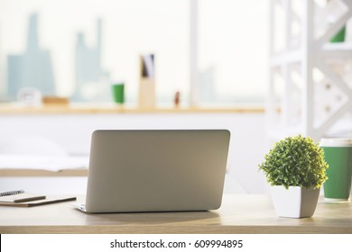 Back View Of Open Laptop Placed On Wooden Desktop With Decorative Plant, Coffee Cup, Supplies And Other Items. Blurry City View Background. Copy Space