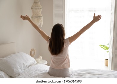 Back View Of Optimistic Young Woman Sit On Bed In Bedroom Wake Up Stretch Or Do Gymnastics. Happy Female Awaken On Early Weekend Morning At Home Feel Overjoyed After Good Sleep. Peace Concept.