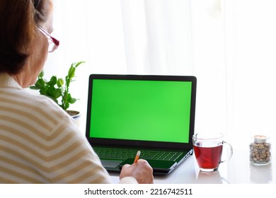 Back View Over Shoulder View Of Senior Elderly Woman With Glasses Watching Educational Webinar, Online Lecture Seminar On Laptop With Green Screen, Taking Notes. The Concept Of Distance Education