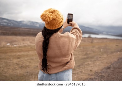 back view overweight model makes photo on phone. Beautiful woman is wearing jeans, sweater and hat walking in cold weather along the road  against the background of landscape and nature. 