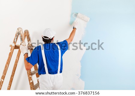Back view of  painter with cap and gloves painting a wall with paint roller and wooden vintage ladder, with copy space