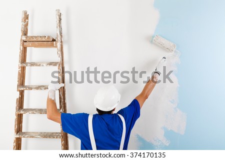 Back view of  painter in white dungarees, helmet and gloves painting a wall with paint roller and wooden vintage ladder, with copy space