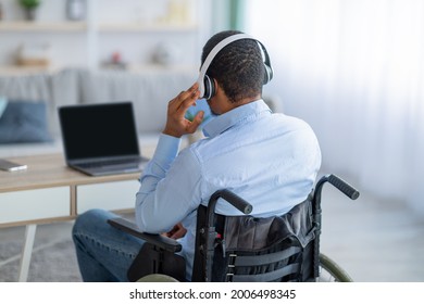 Back View Of Paraplegic Black Guy In Wheelchair Wearing Headphones, Using Laptop With Mockup For Design At Home. New Normal, Be Safe, Remote Job, Online Education, Study And Freelance Concept