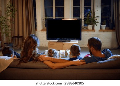 Back view of parents and children spending evening together, sitting on couch in living room and watching tv, eating snacks. Concept of family, leisure time, relaxation, childhood and parenthood - Powered by Shutterstock