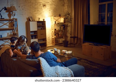 Back view of parents and children spending evening together, sitting on couch in living room and watching tv, eating snacks. Concept of family, leisure time, relaxation, childhood and parenthood - Powered by Shutterstock