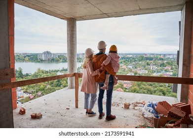 Back View Of Parents With Daughter Standing Inside Apartment Building Under Construction. Man And Woman With Child Wearing Building Helmets While Enjoying City View From Future Home.