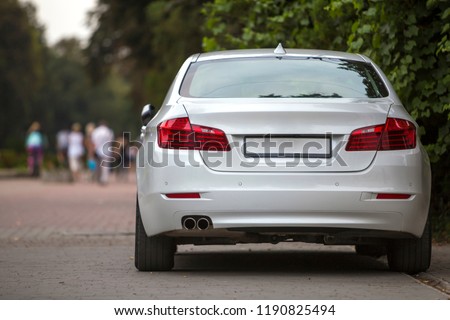 Back view part of white car parked on city pedestrian zone pavement on background of blurred silhouettes of people walking along green sunny summer alley. Modern city lifestyle concept.