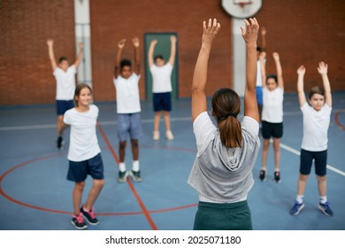 Back View Of PE Teacher And Group Of Elementary Students Exercising During A Class At School Gym. 