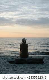 Back view of pensive lonely woman in coat and hat sitting on log on seashore looking away at sea in evening.