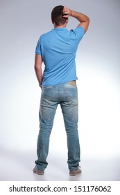 Back View Of A Pensive Young Casual Man Scratching His Head While Holding A Hand In His Pocket. On Gray Background