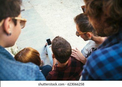 Back View Of People Sitting Together Outdoors And Watching Smartphone.