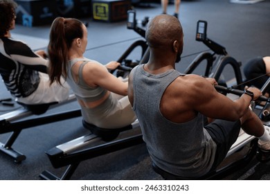 Back view of people using rowing machine during strength training in gym with muscular man in foreground - Powered by Shutterstock