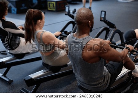 Back view of people using rowing machine during strength training in gym with muscular man in foreground