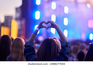 Back view of person forming heart shape with hands at music fest, surrounded by cheering crowd, vibrant stage lights blur in background, festive event atmosphere captures moment of joy at live concert