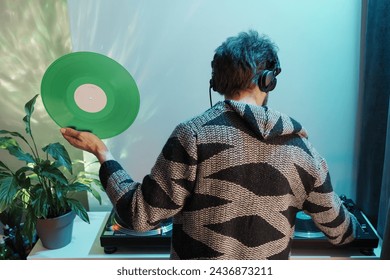 Back view of a person with headphones holding a vinyl record, standing by a turntable and indoor plants. - Powered by Shutterstock