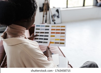 Back View Photo Of Young African Woman Interior Designer Sitting On Sofa Indoors At Home While Holding Catalogue Of Colors